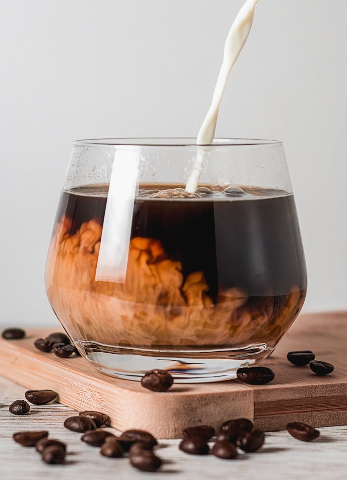 Close-up of creamy milk pouring into a glass of coffee surrounded by coffee beans on wooden boards.
